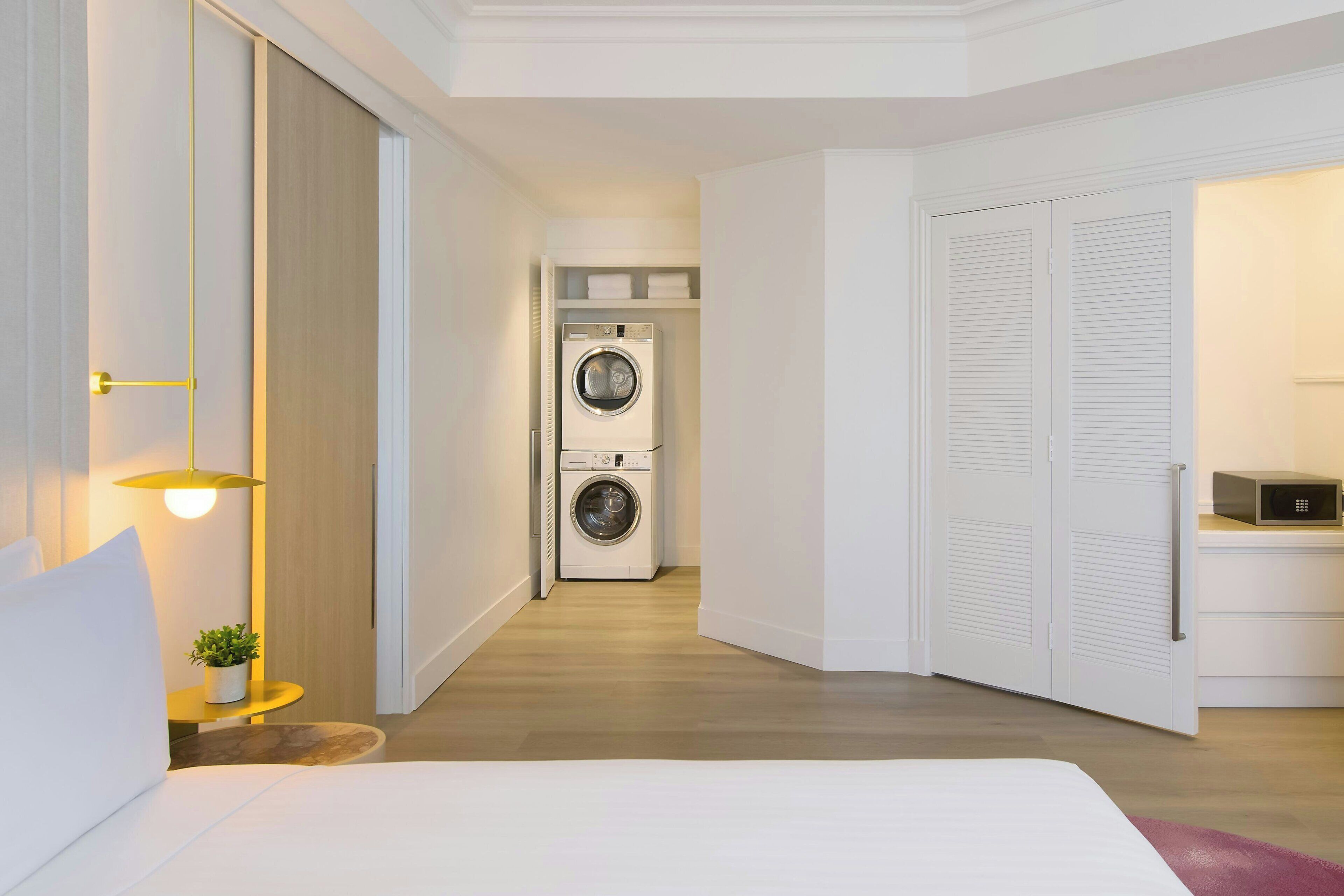A hotel room with a bed, a washing machine, a small table with a plant, and a lamp.