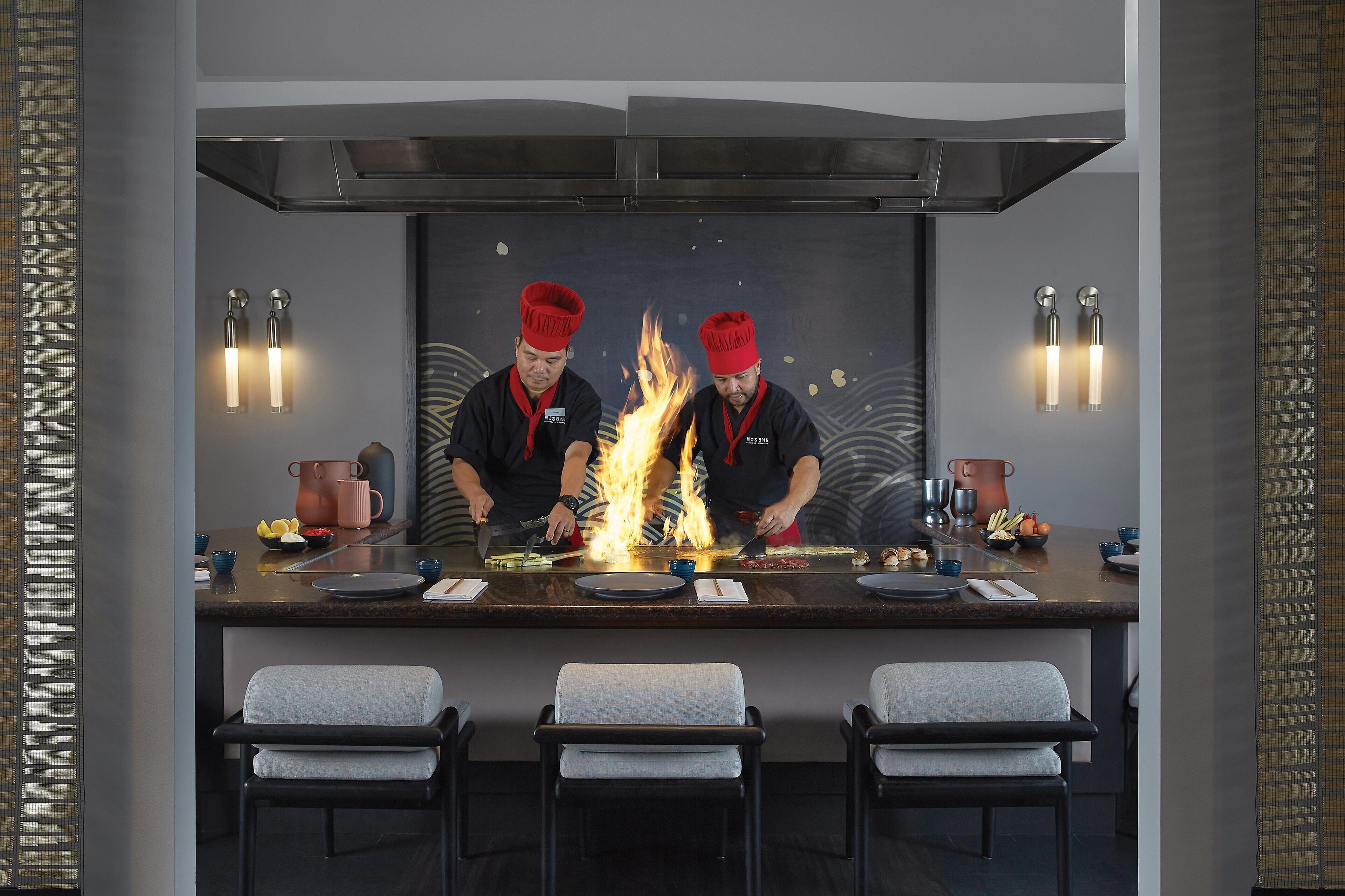 A modern kitchen with chefs preparing food over an open flame, a bar area with seating, and contemporary decor.