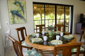 A dining area with a round table set for four, surrounded by wooden chairs. There is a painting of birds on the wall, a thatched roof visible through the window, and a television on a stand to the right.