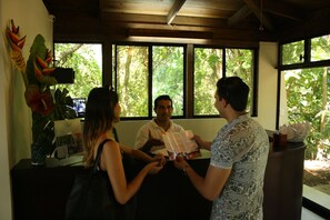 A reception area with a wooden ceiling, large windows with a view of greenery, and a reception desk with a plant and a decorative item.