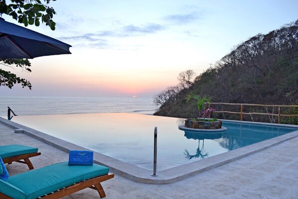 An infinity pool with a view of the ocean at sunset, lounge chairs, and a small island with plants.