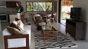A living room with a sofa, coffee table, and TV stand. There is a dining area with a table and chairs. - Paraiso Escondido Hotel Villas & Resort (Teotepeque)
