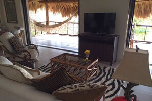 A living room with a sofa, armchairs, a glass coffee table, a TV, and a hammock outside.