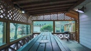 Ein überdachter Außenbereich mit Holztisch und -stühlen, einer Gitterwand und großen Fenstern.