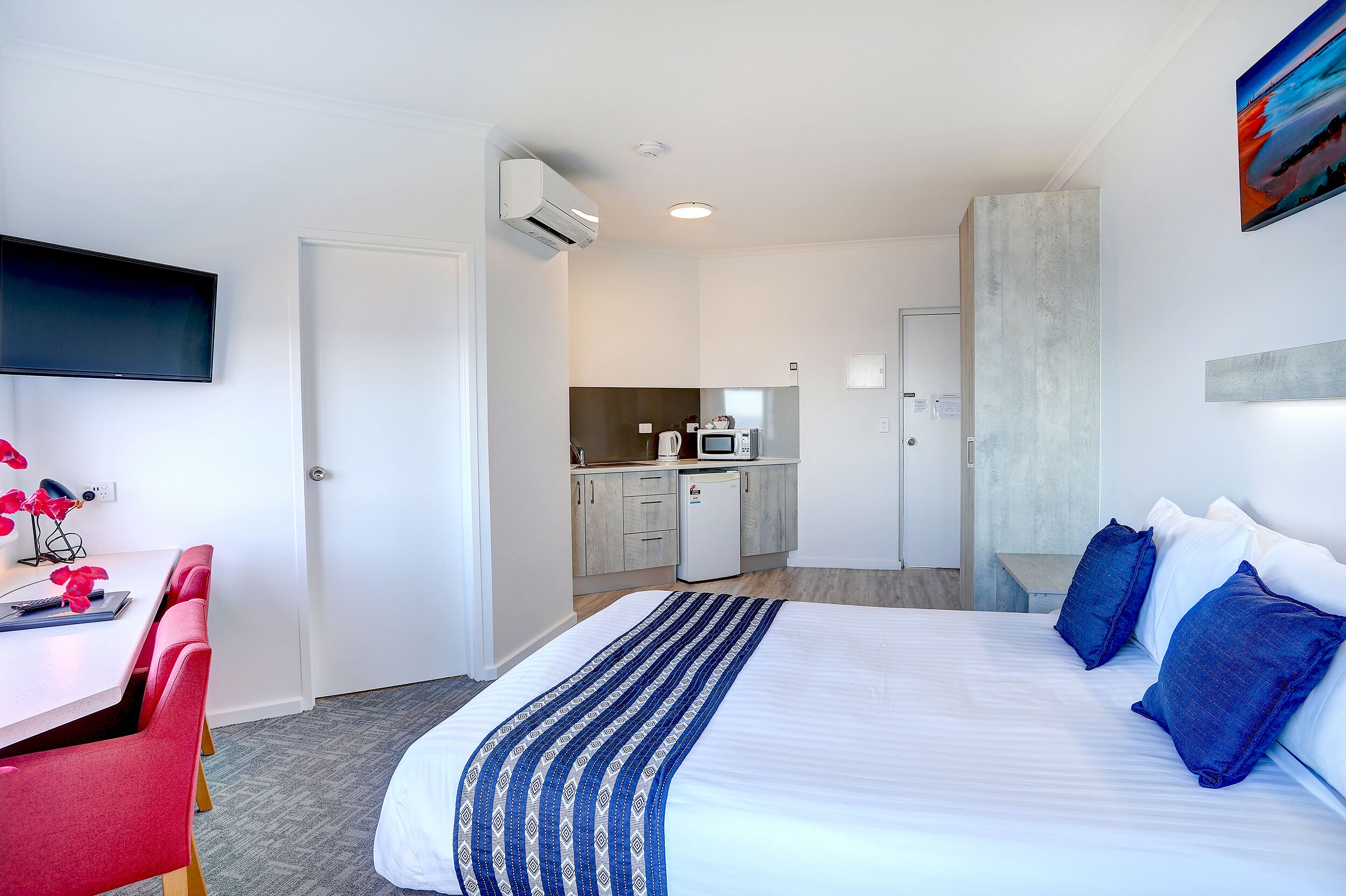 A hotel room with a bed, a desk with a chair, a kitchenette, and a TV mounted on the wall.