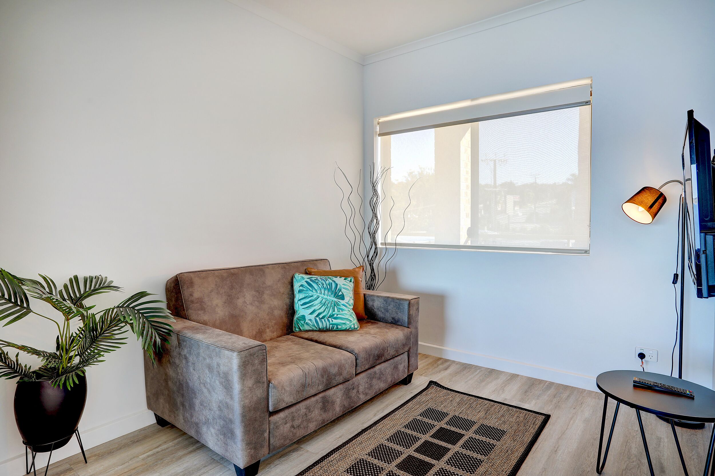 A modern living room with a brown sofa, a large window, a floor lamp, and a potted plant.