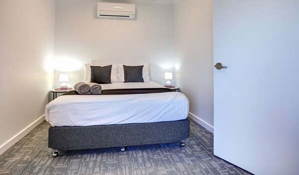 Una cama bien hecha con ropa de cama blanca y gris oscuro, dos lámparas de noche y un aire acondicionado instalado en la pared.