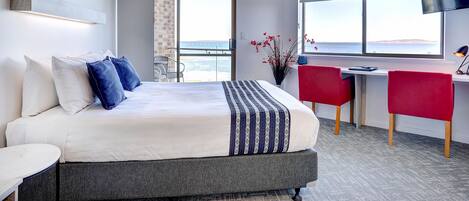 A modern hotel room with a large bed, a desk with two red chairs, a TV, and a view of the sea.