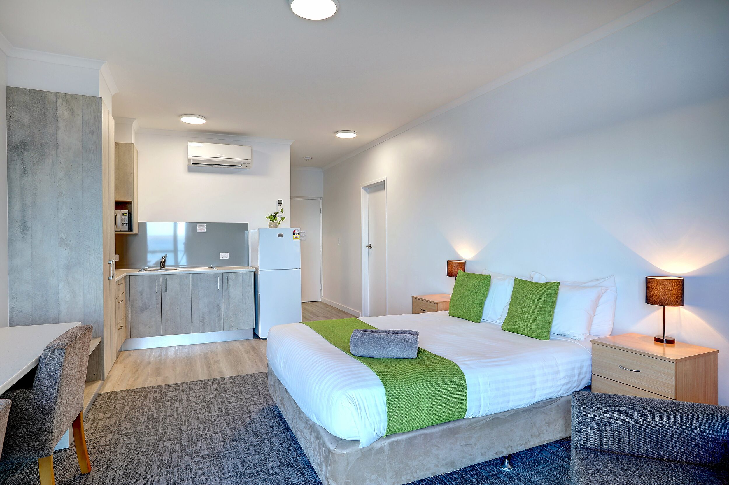 A modern hotel room with a bed, bedside tables, a desk, and a kitchenette.