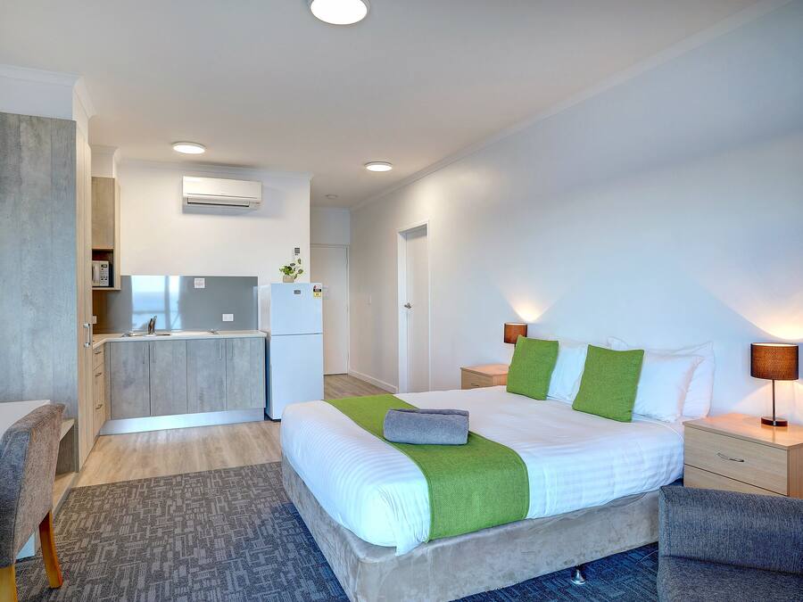 A modern hotel room with a bed, bedside tables, a desk, and a kitchenette.