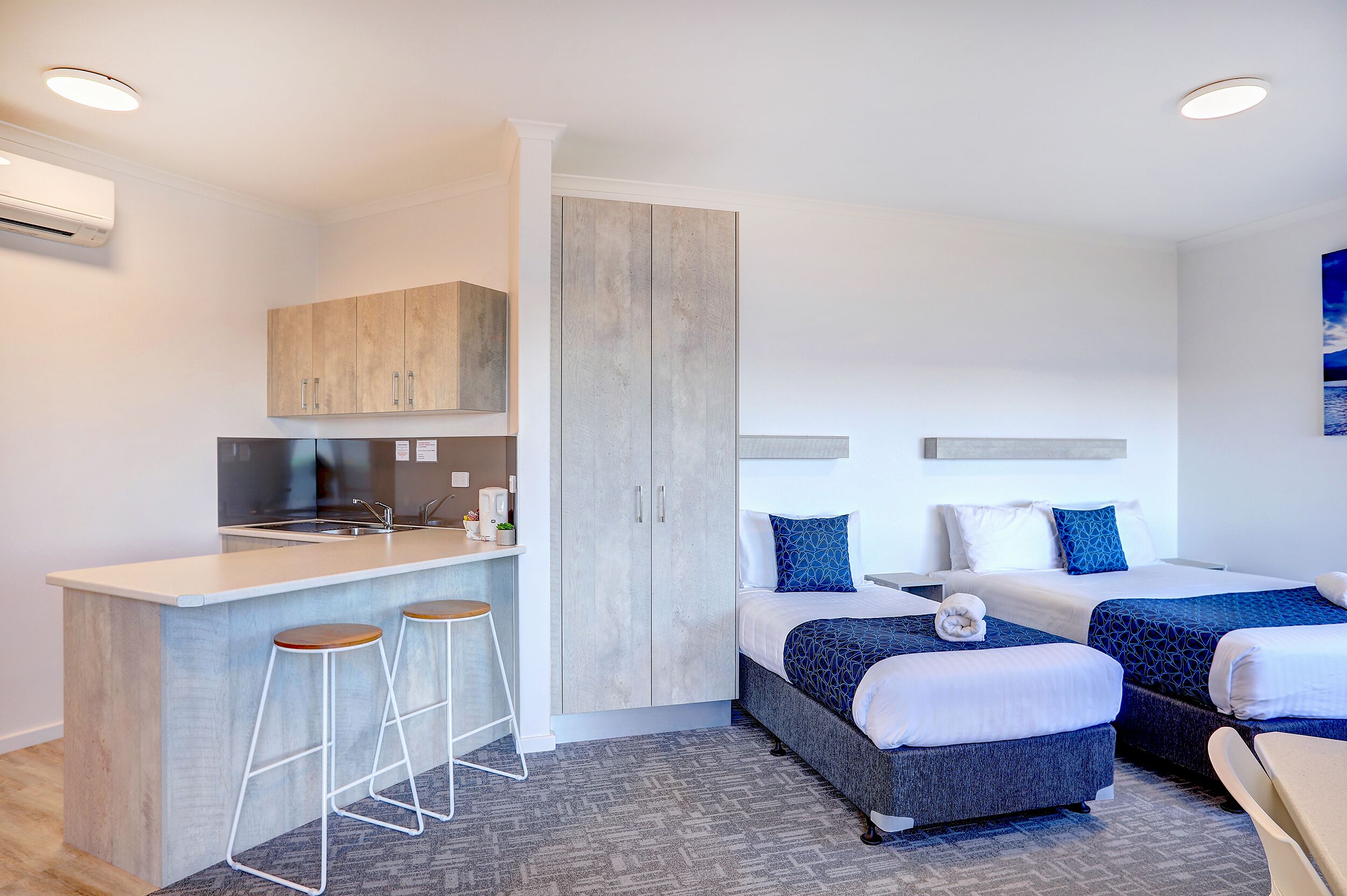 Une chambre d’hôtel moderne équipée d’une kitchenette, de deux lits, d’un tabouret de bar et d’une grande armoire.