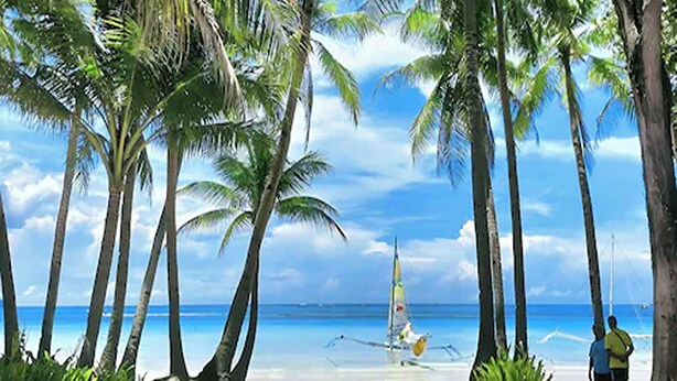 A beach scene with palm trees, a clear sky, and a sailboat in the distance. - Zuzuni Boutique Hotel (Boracay Island)