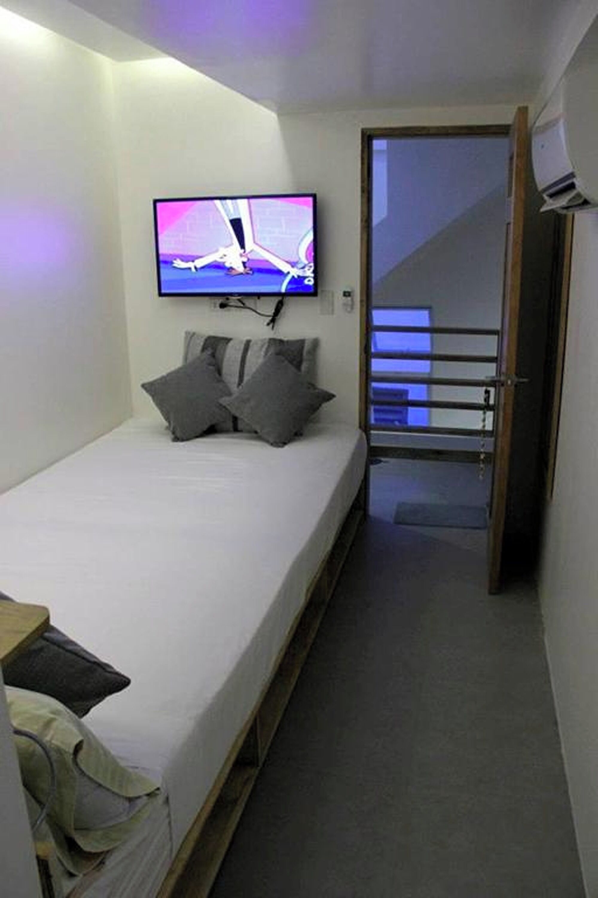 Une petite pièce étroite, avec un lit simple, un téléviseur fixé au mur et une porte donnant sur un escalier.