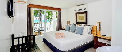 Quarto de hotel com uma cama grande, um banco, uma televisão, uma mesinha de cabeceira de madeira e vista para a praia.