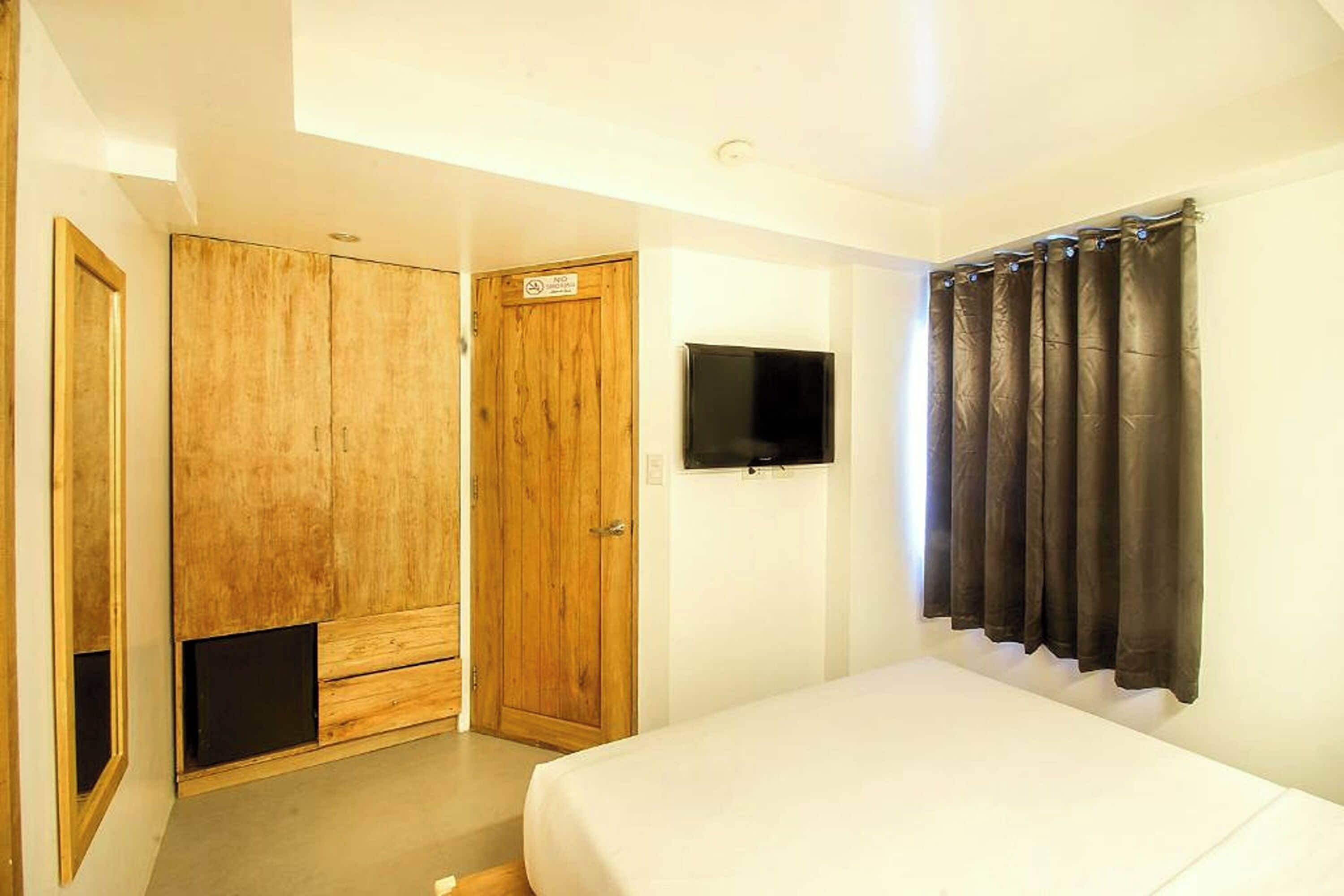 Une chambre à coucher avec un lit, une armoire en bois, une télévision et une fenêtre avec des rideaux.
