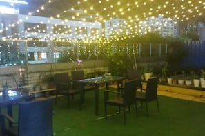 An outdoor seating area with tables and chairs, string lights overhead, and a view of buildings in the background.