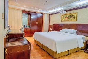 A hotel room with a bed, wooden desk, wardrobe, and a painting on the wall.