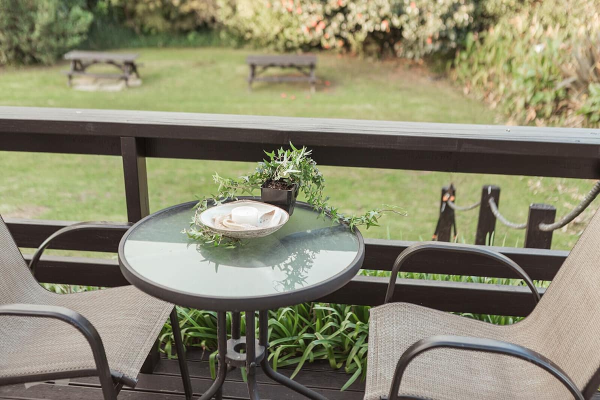 A glass-top table with a bowl of cream cheese and crackers, surrounded by two chairs, on an outdoor patio with a view of a garden and picnic tables in the background.
