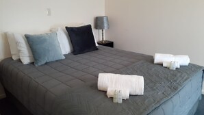A neatly made bed with a grey quilt, multiple pillows in white, blue, and black, a bedside lamp, and two towels and bottles on the bed. - Golden Bay Motel (Takaka)