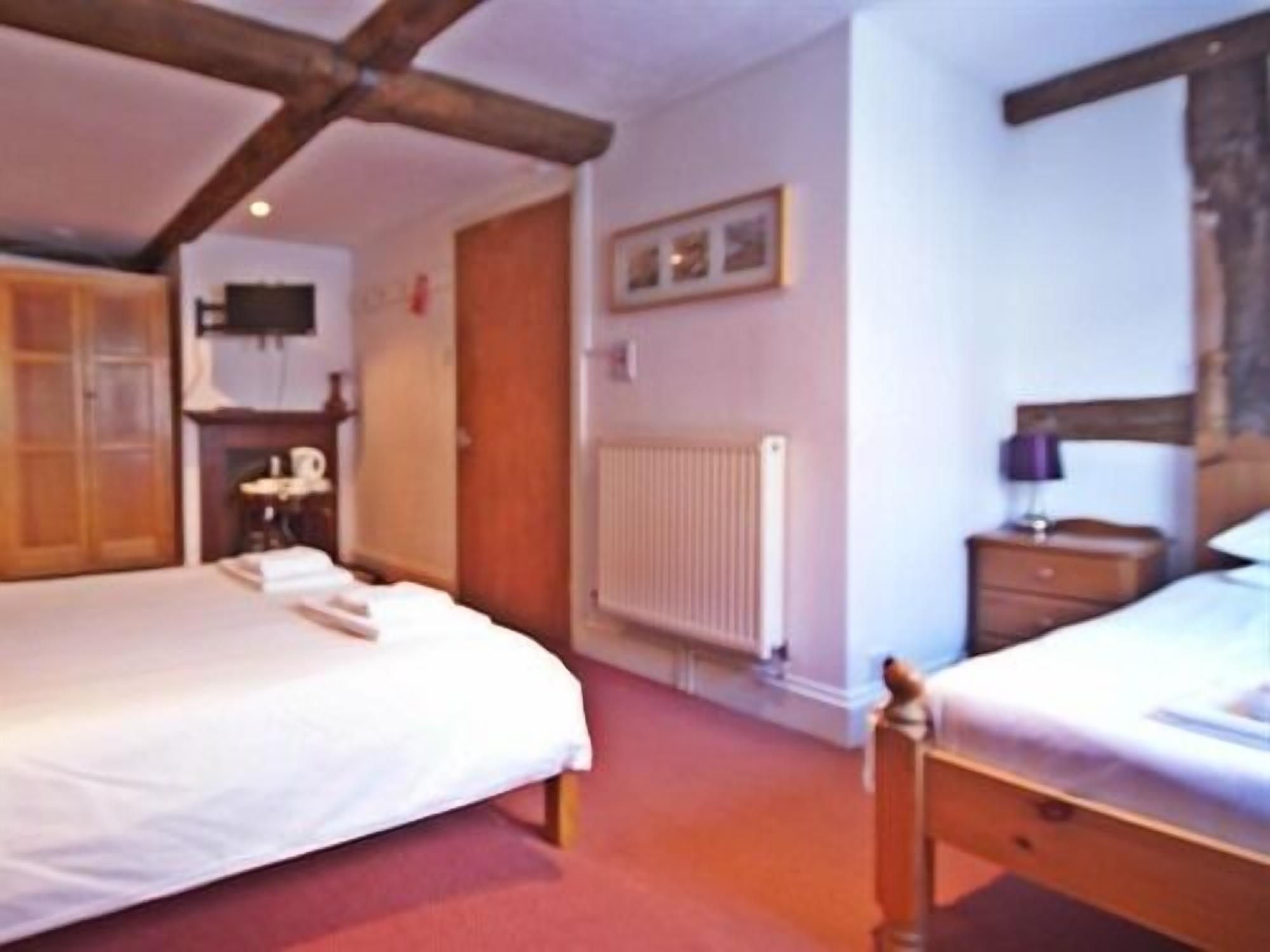 A bedroom with two beds, wooden beams on the ceiling, a radiator, and a nightstand with a lamp.
