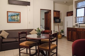 A living room with a brown sofa, a dining table with chairs, a kitchen area with wooden cabinets, and a painting on the wall. - Hotel Plaza Real (San José)