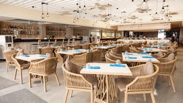 Un restaurante espacioso con sillas y mesas de mimbre, una zona de bar con diversas botellas, y un diseño de techo moderno.