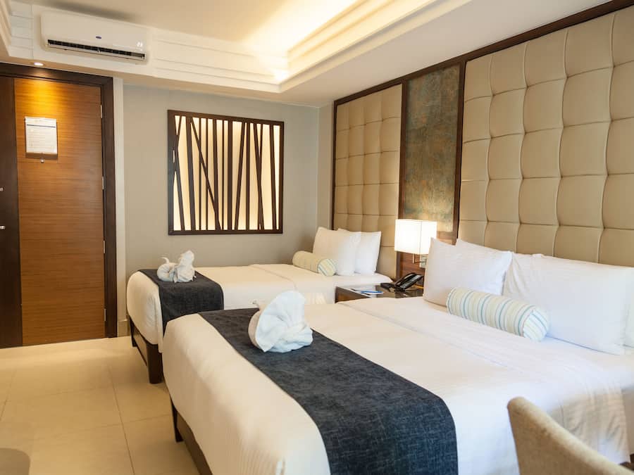 A hotel room with two beds, a wooden door, a wall-mounted air conditioner, a decorative panel, and a bedside lamp.