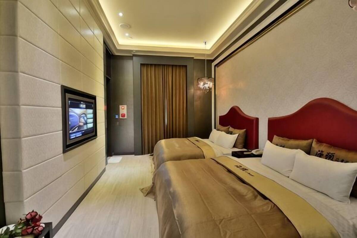 Quarto de hotel com duas camas, uma televisão fixada na parede e um lustre moderno.