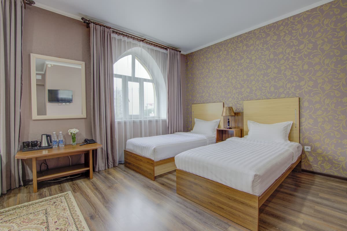 Hotellihuone, jossa on kaksi erillistä sänkyä, puinen työpöytä, peili, ikkuna, jossa on verhot, ja kuviollinen tapetti.