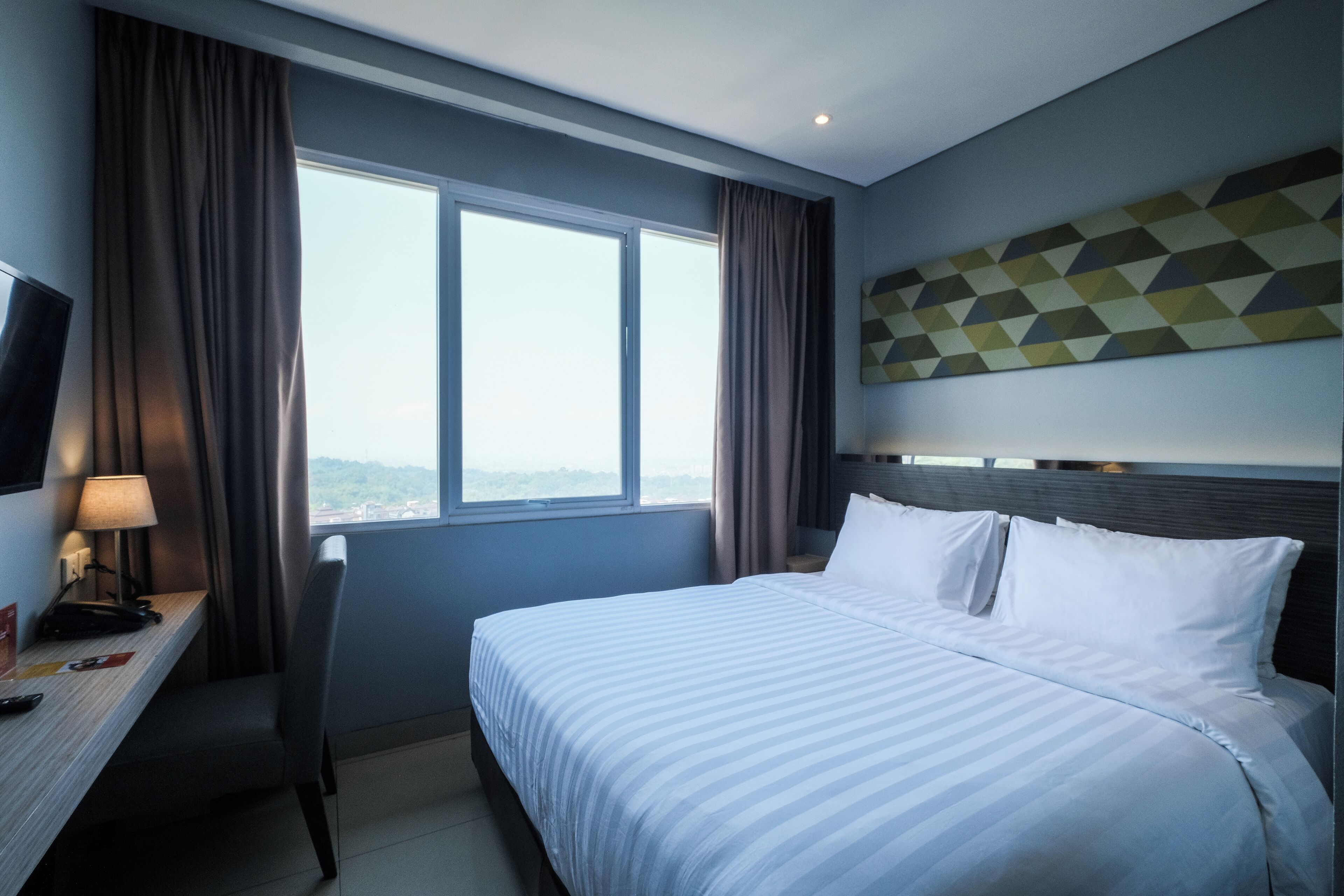 A hotel room with a bed, a desk with a lamp, a TV, and a window with curtains.