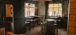A cozy cafe with wooden tables and chairs, large windows with natural light, and a brick wall with photos. - The Crafty Cow (Bristol)