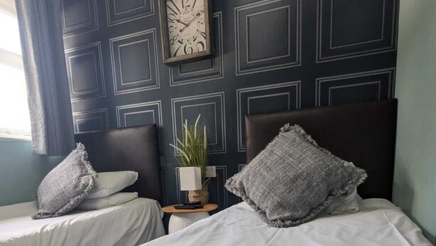Une chambre d’hôtel avec deux lits, un lambris sombre, une horloge, une petite table avec une plante et une serviette pliée posée sur le lit.
