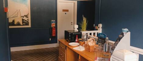 A hotel room with a wooden desk, a microwave, a fire extinguisher, a plant, and a sign for 'Navas'.