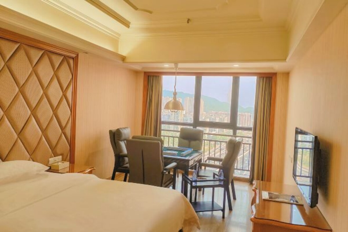 Una habitació d'hotel amb un llit gran, una taula de treball amb cadires, una televisió i vista a la ciutat a través de grans finestres.