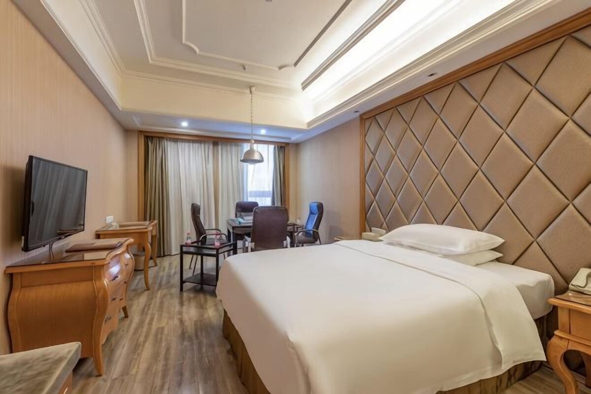 Een hotelkamer met een groot bed, een bureau met stoel, een televisie en een raam met gordijnen.