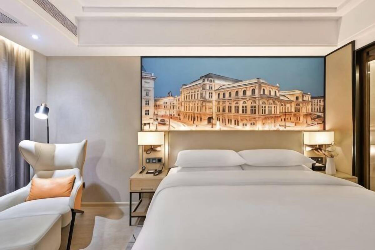 A hotel room with a large bed, a desk, a chair, and a view of a building through a window.