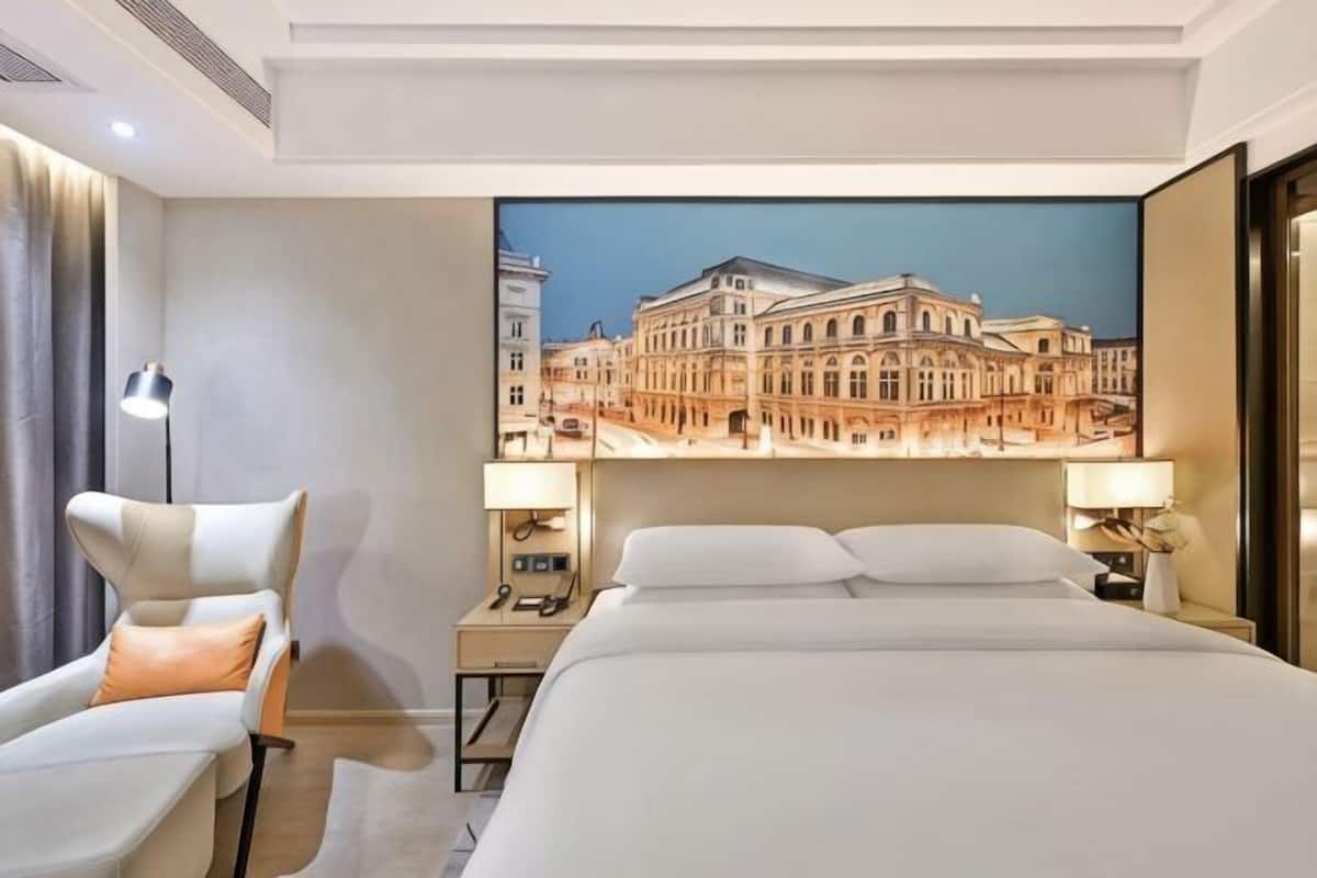 A hotel room with a large bed, a desk, a chair, and a view of a building through a window.