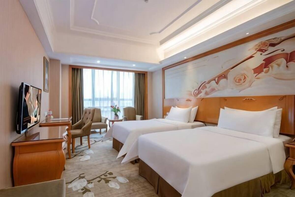 A hotel room with two beds, a desk, a chair, a TV, and a large window.