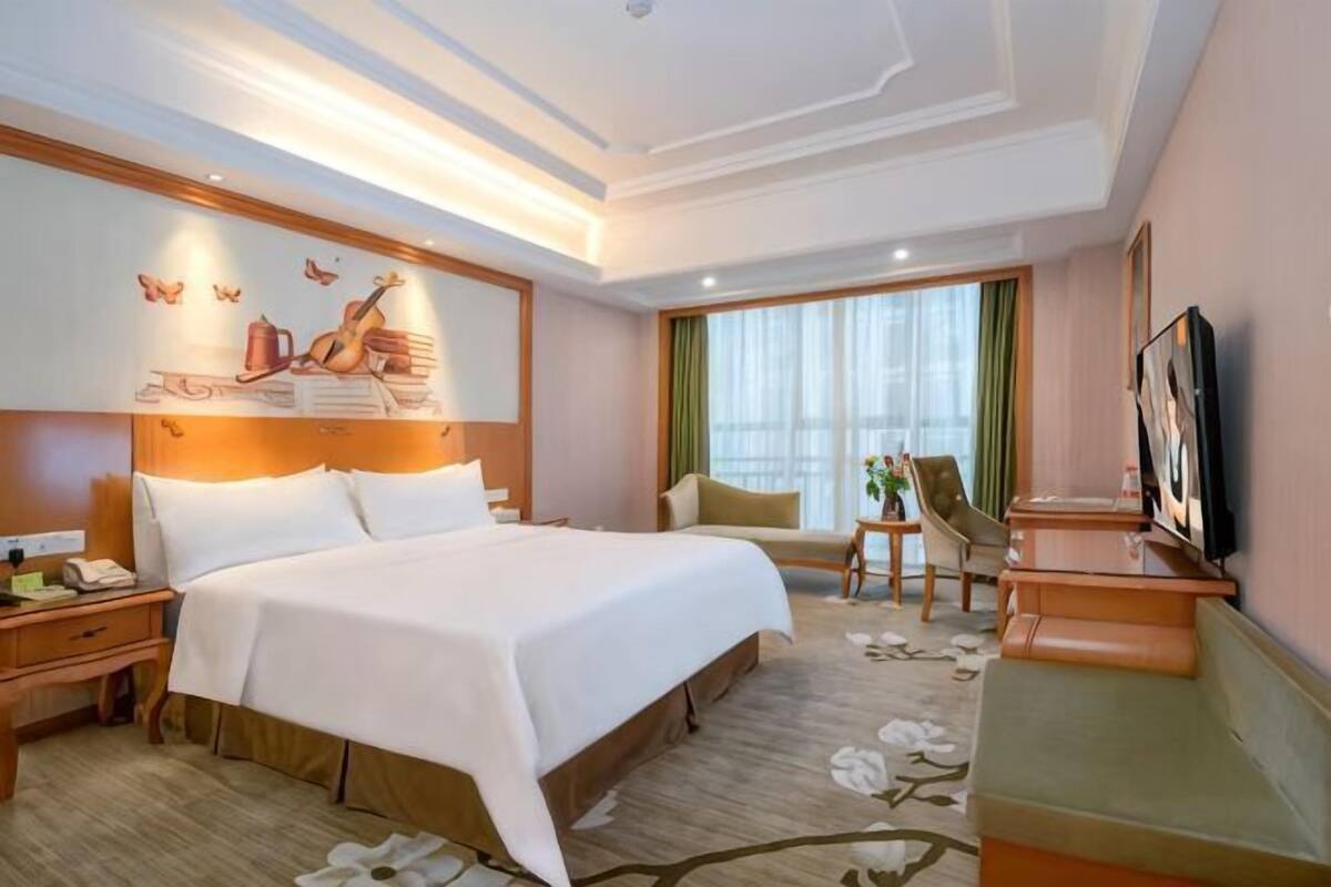 A hotel room with a large bed, bedside tables, a desk, a chair, a TV, and a window with curtains.