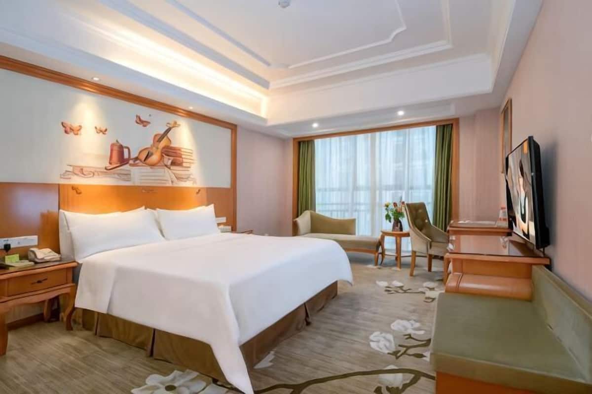 A hotel room with a large bed, bedside tables, a desk, a chair, a TV, and a window with curtains.