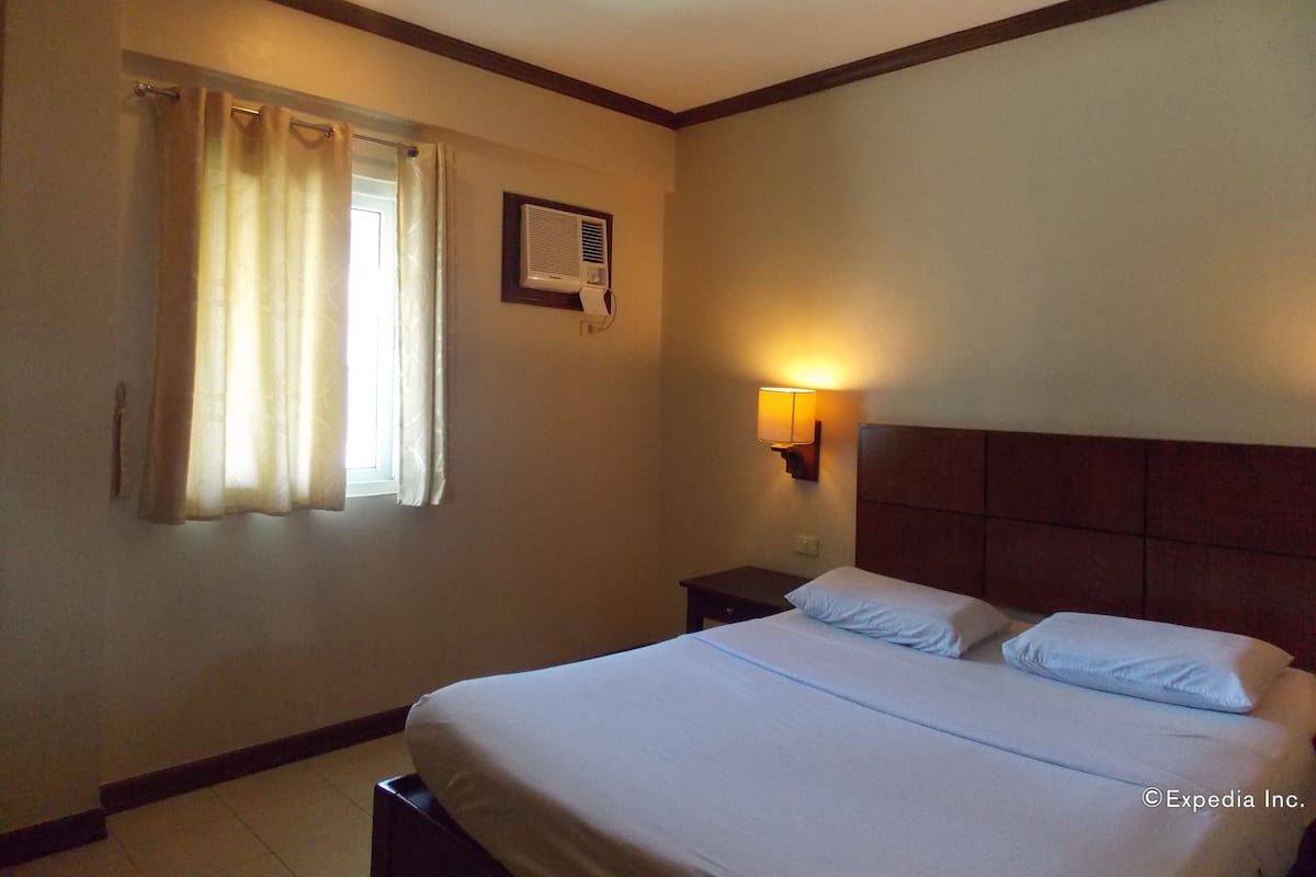 Una habitación de hotel con cama, mesita de noche, aire acondicionado instalado en la pared y ventana con cortinas.