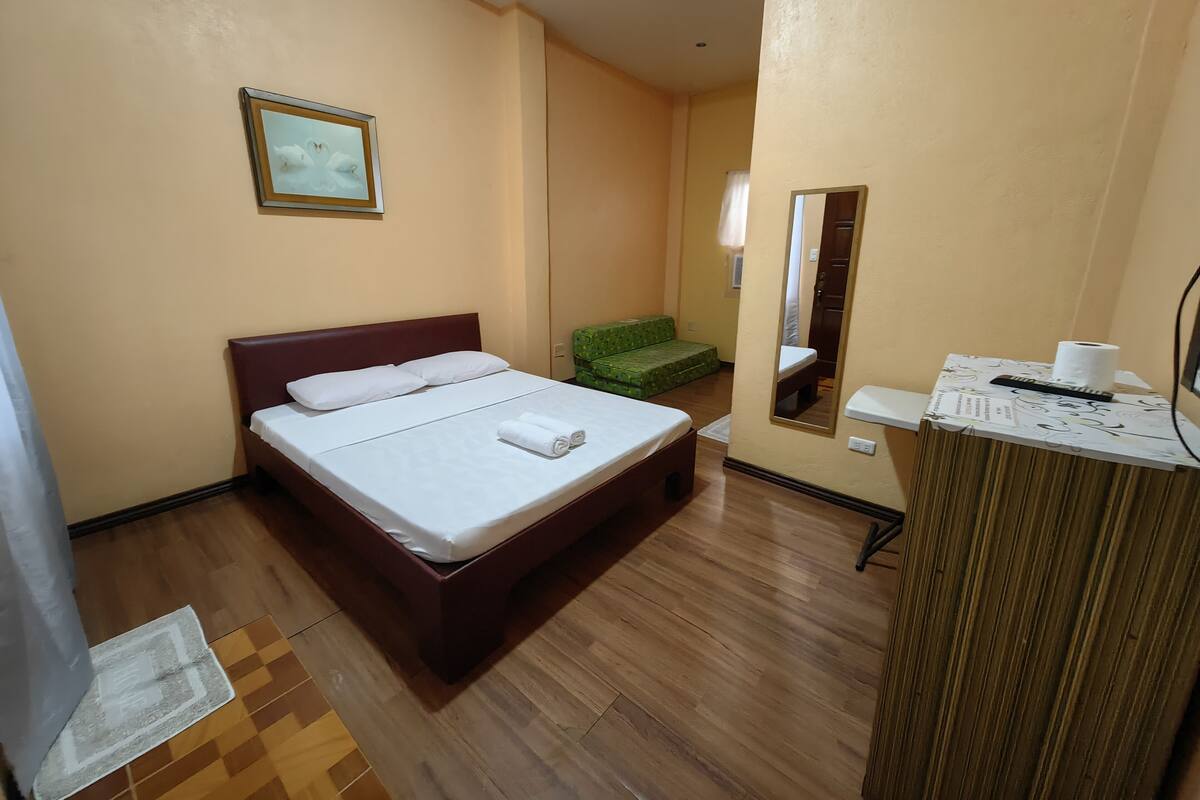 A hotel room with a bed, a small green sofa, a mirror, and a desk with a toilet paper roll.