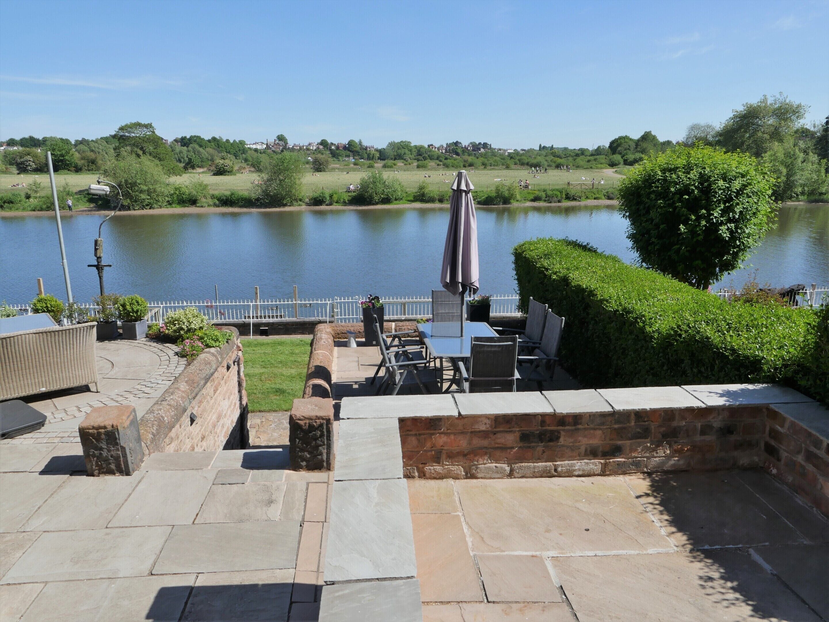 A patio area with stone flooring, outdoor furniture, and a view of a lake with a white fence and greenery in the background.