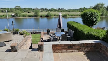 A patio area with stone flooring, outdoor furniture, and a view of a lake with a white fence and greenery in the background.