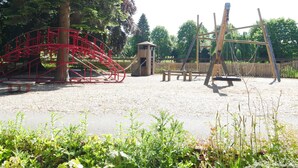 A playground with a red climbing structure, swings, and a small wooden shelter. - Rondeva (Chester)