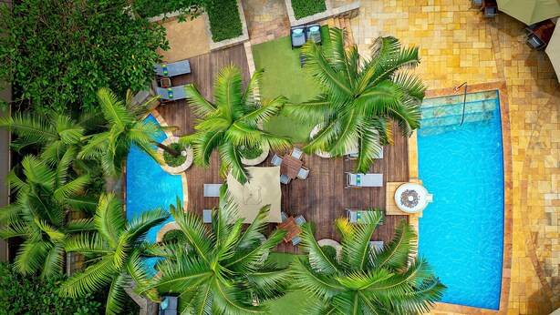 Eine Luftaufnahme eines Resorts mit einem zentralen Swimmingpool, umgeben von Palmen und Sitzbereichen im Freien.