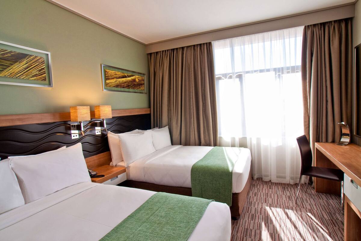 Une chambre d’hôtel avec deux lits, un bureau, une chaise et une grande fenêtre avec des rideaux.