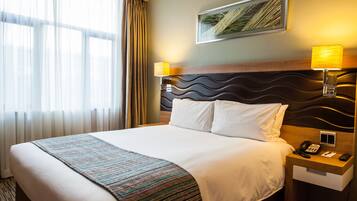 Una habitación de hotel con una cama grande, dos lámparas de noche, una mesita de noche con un teléfono y un control remoto, y una ventana con cortinas.