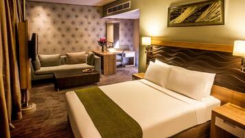 Habitación de hotel con una cama grande, un sofá, un escritorio y una silla. Se observa un espejo, una lámpara y un cuadro en la pared.