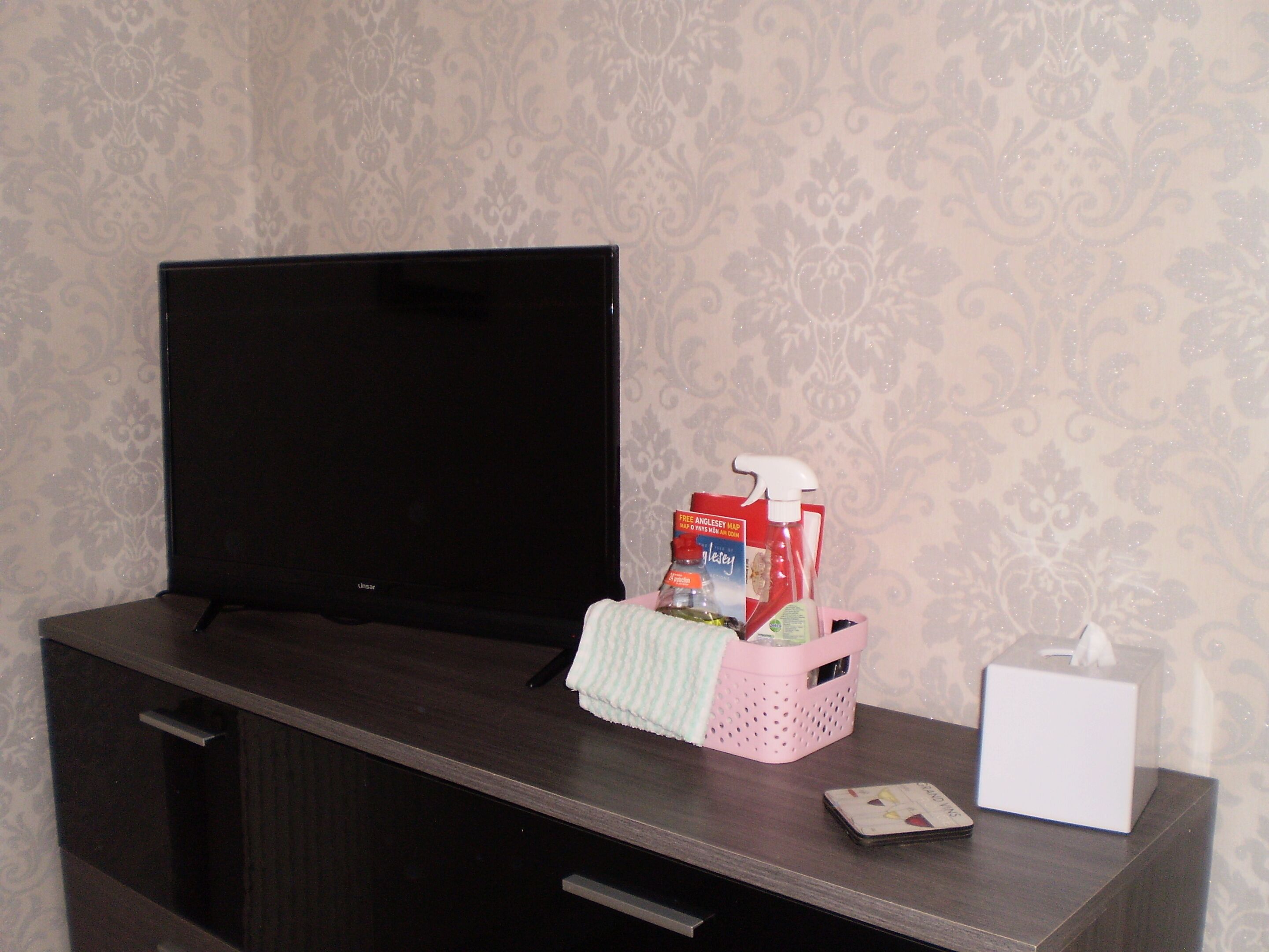 Een donker houten bureau met een televisie, een roze mand met schoonmaakspullen en een zakdoosje.